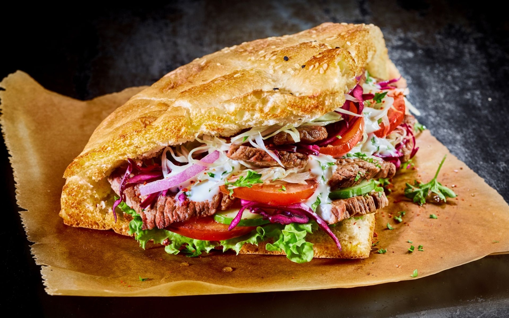 Ein großes, gefülltes Sandwich mit Roastbeef, Tomaten und grünem Salat, präsentiert auf Backpapier.