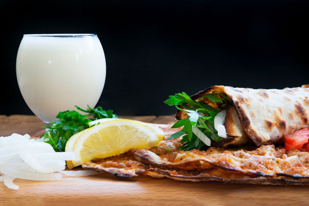 Ein Frühstücksarrangement mit einem gefüllten Wrap, einem Glas Milch, Zitronenscheiben und frischem Gemüse auf einem Holzbrett.